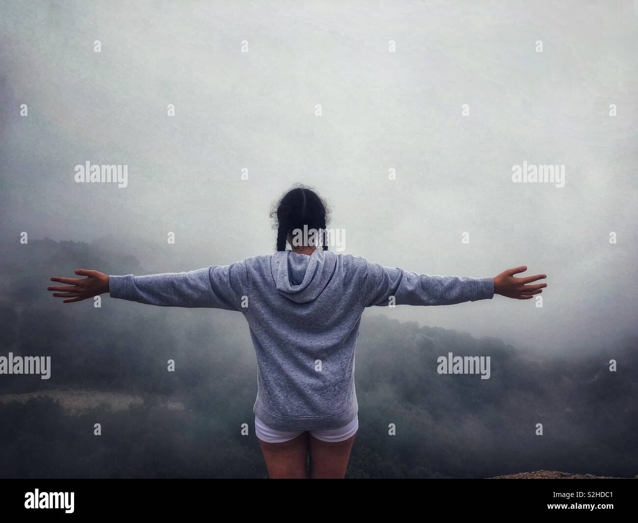 Das Mädchen steht, die Arme ausgestreckt, auf einem Berg mit Nebel aus einer absteigenden Wolke bedeckte. Stockfoto