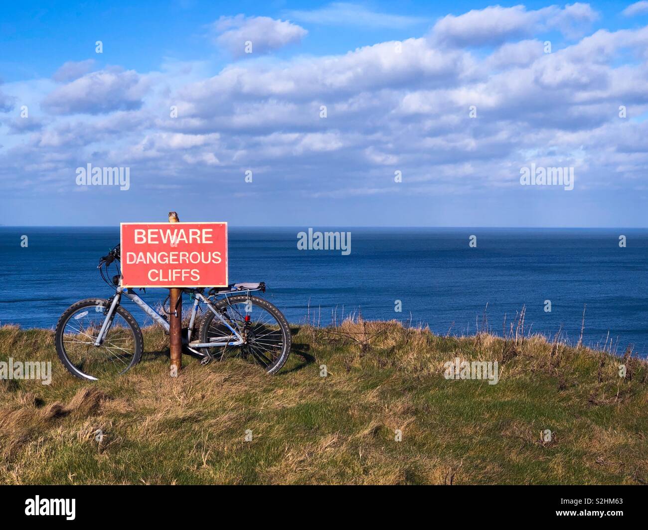 Fahrrad gebunden an eine Warnung Wegweiser auf den Rand einer hohen Klippe mit Blick auf das Meer an einem sonnigen Tag. Stockfoto