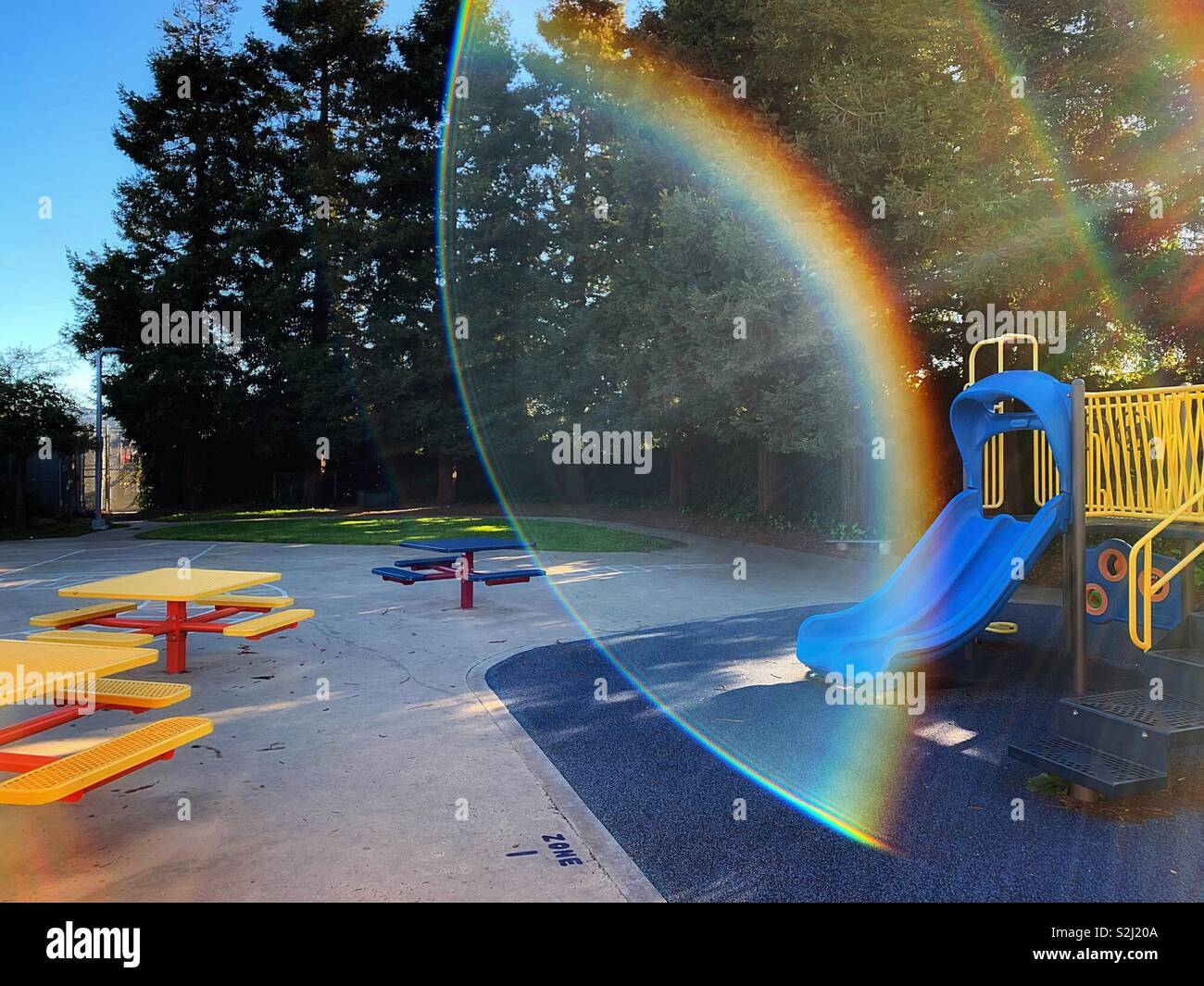 Die gelben und blauen Spielplatz Struktur, die gelbe Tische mit Bänken, die riesigen roten Wald Bäume und so weiter in den Kinderspielplatz Szene über den blauen Himmel Stockfoto