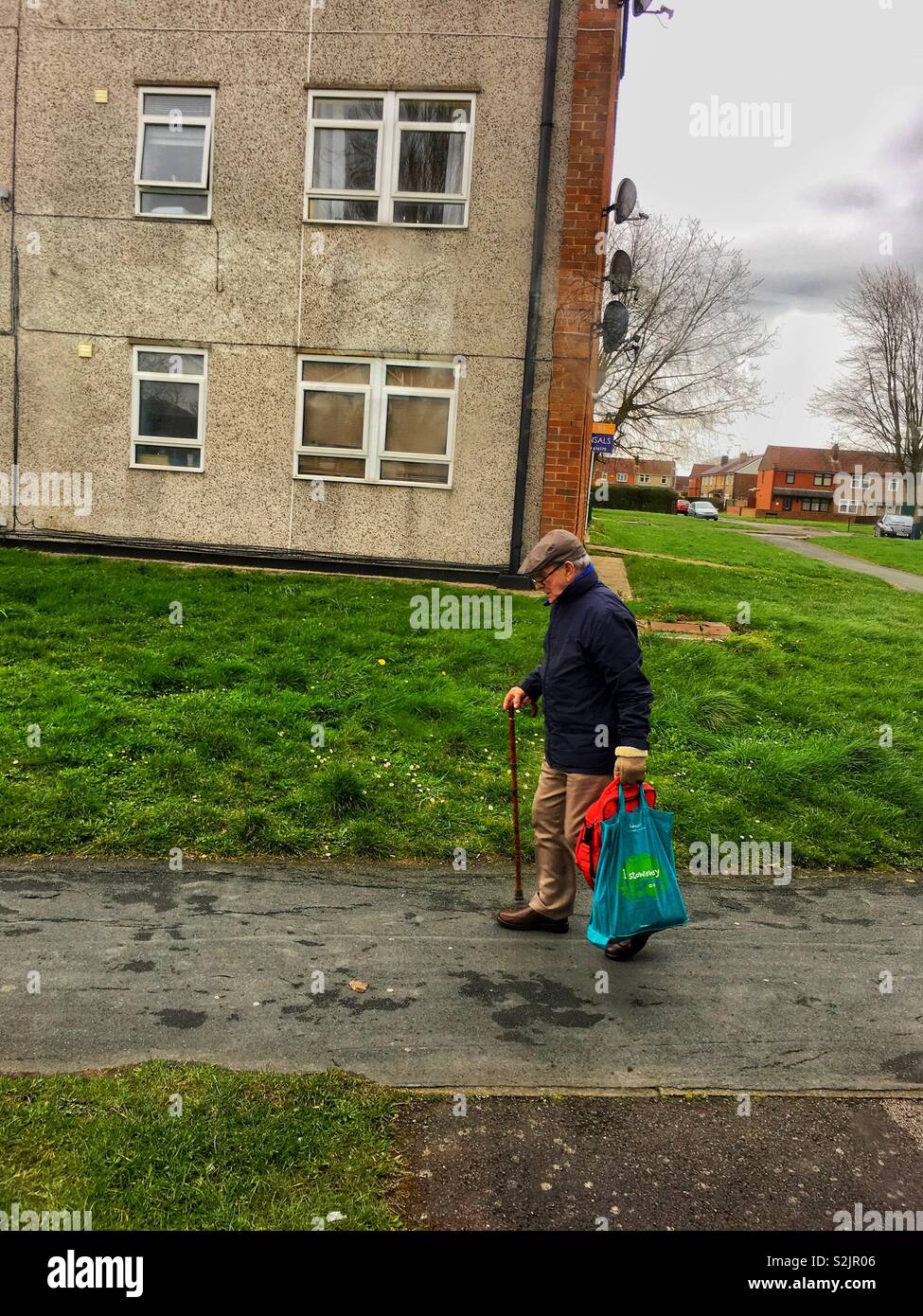 Ein älterer Mann in einer flachen Kappe, mit einem Stock und Kunststoff Einkaufstaschen, Spaziergänge entlang der Fahrbahn außerhalb eines Mehrfamilienhauses. Platz kopieren Stockfoto