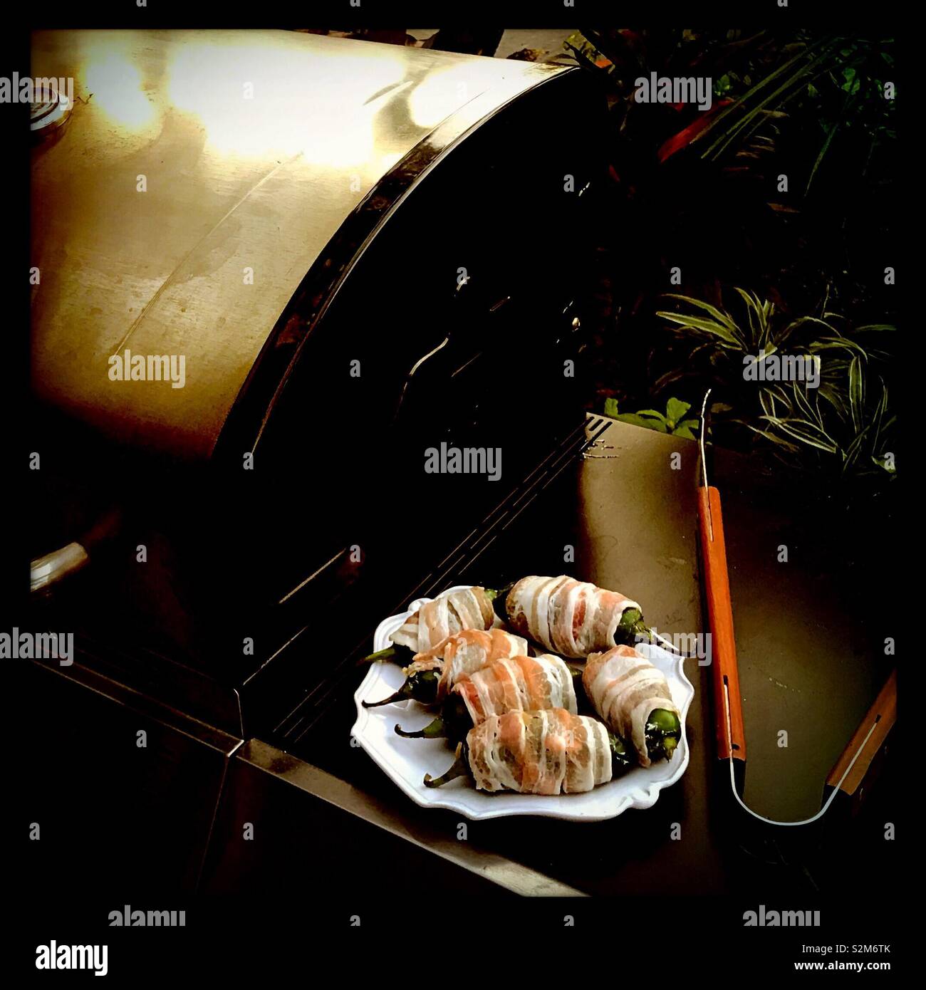 Eine Platte mit Speck umwickelt jalapeños sitzt auf dem Grill Leiste neben einigen Zangen als Grill wärmt für das Grillen. Stockfoto