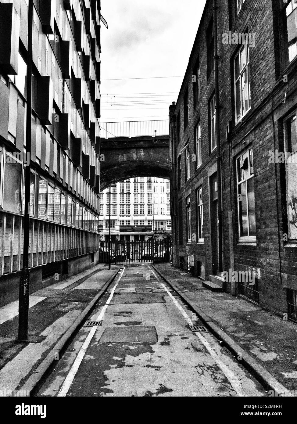 Ein Schwarzweißfoto einer engen Gasse in Manchester, flankiert von modernen und alten Gebäuden, mit einer Überführung im Hintergrund - Smartphone-aufgenommenes Stockfoto