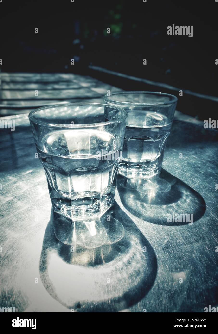Zwei teilweise volle Gläser von Wasser auf der sonnenbeschienenen Terrasse Tisch aus Metall mit Lichtbrechung Muster und Schatten. Stockfoto