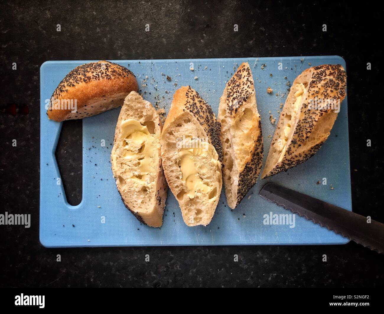 In Scheiben geschnitten und mit Butter bestrichenen Mohn baguette auf einem blauen Schneidebrett. - Smartphone-aufgenommenes Stockfoto