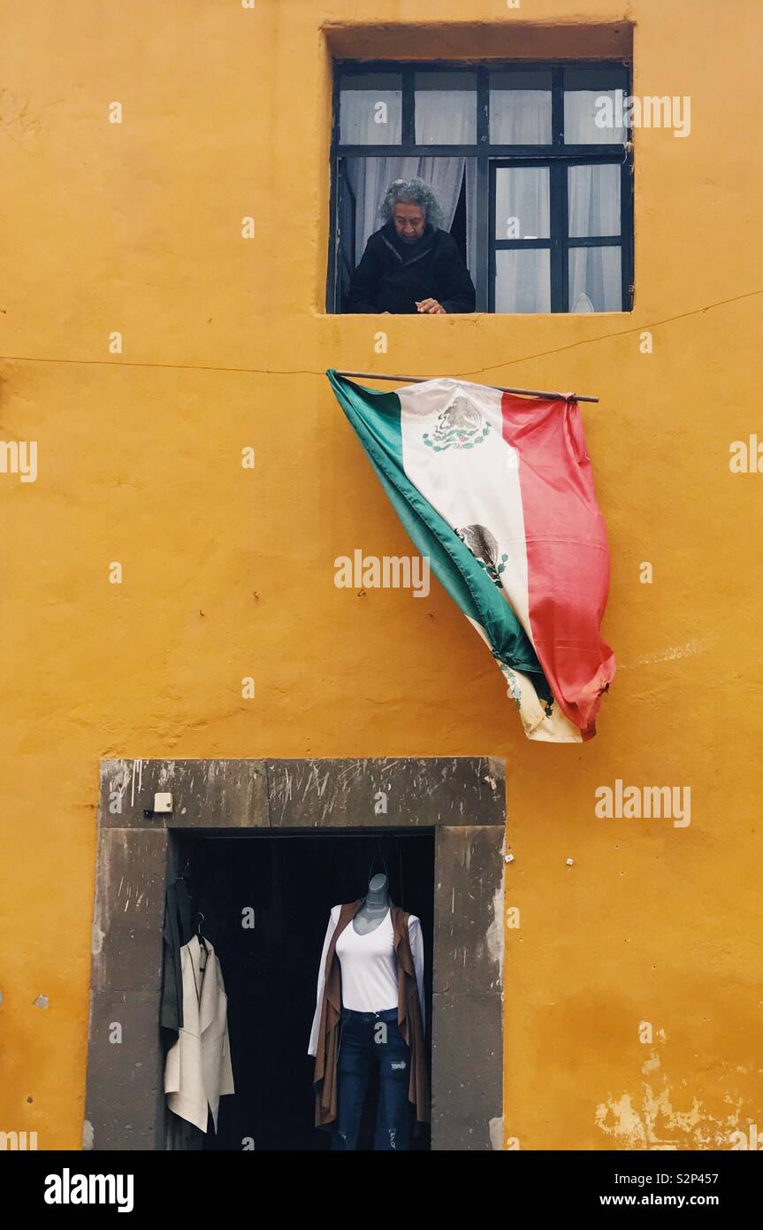 Eine alte Frau schaut man auf der Mexikanischen Flagge von Ihrem Haus oberhalb einer Modeboutique in San Miguel de Allende, Mexiko. Stockfoto