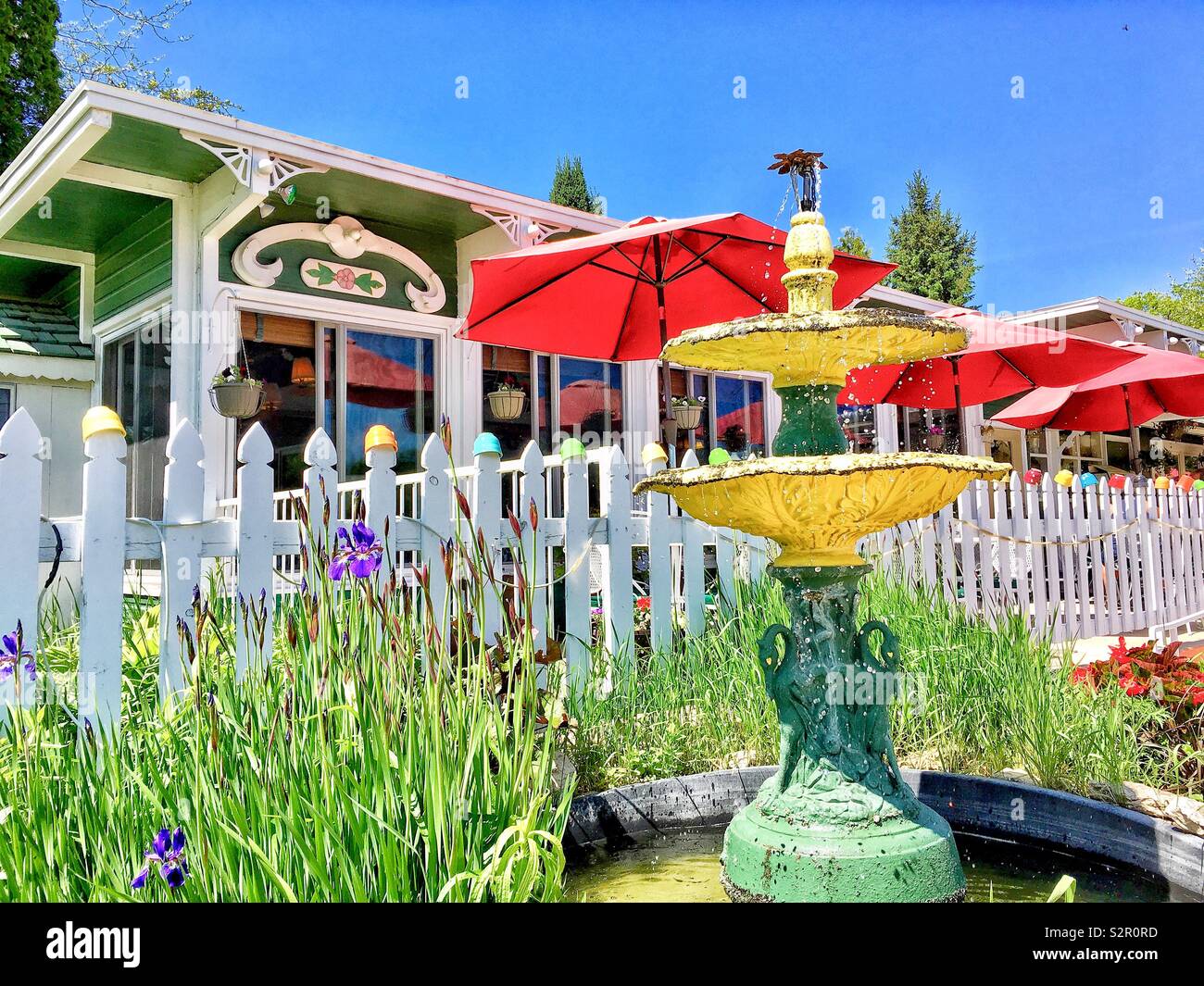 Gelbes Wasser Brunnen mit Bronze Vögel durch Blume Garten von Lila Flieder neben weißen Lattenzaun durch einen Regenbogen von Suppe Becher außerhalb der Sommer Küche Restaurant in Ephriam, Door County gekrönt - Smartphone-aufgenommenes Stockfoto