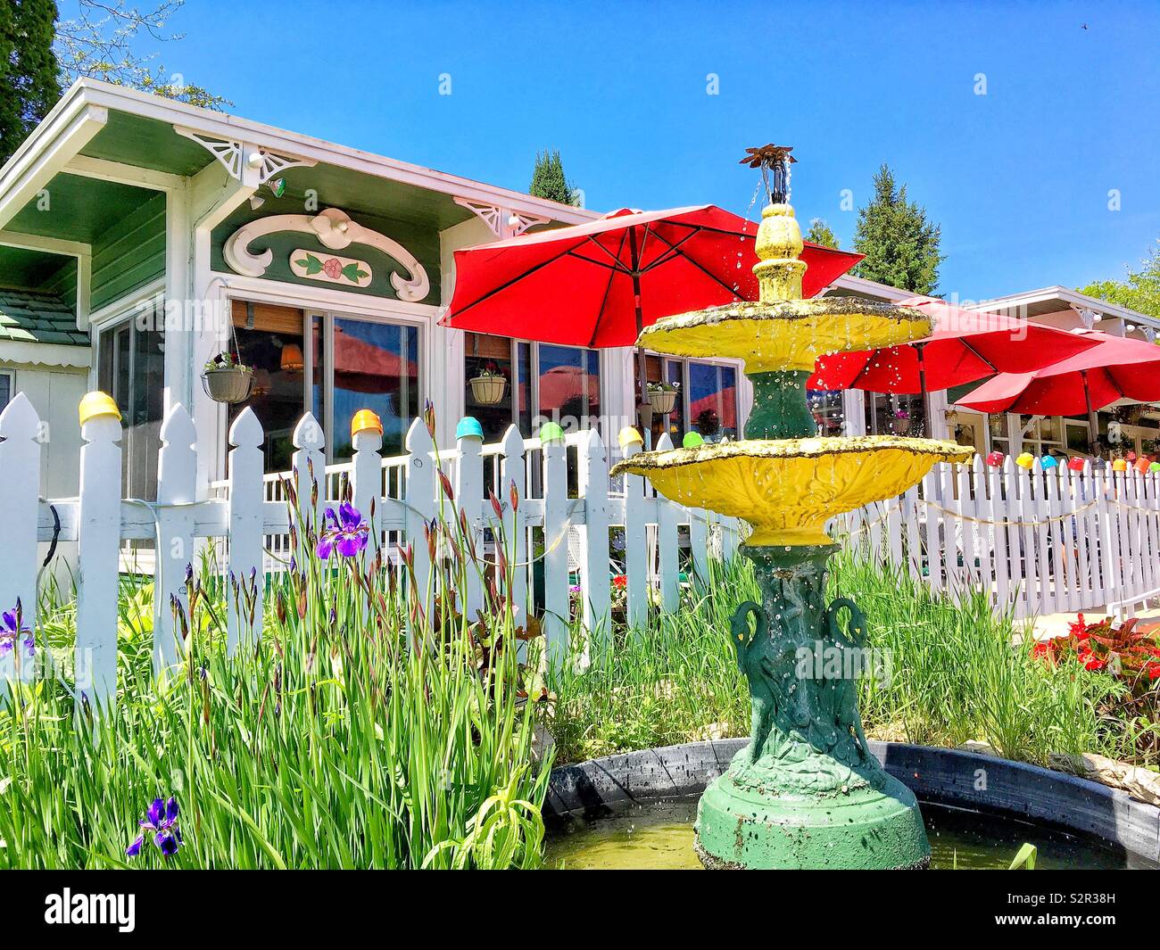Gelbes Wasser Brunnen mit Bronze Vögel durch Blume Garten von Lila Flieder neben weißen Lattenzaun durch einen Regenbogen von Suppe Becher außerhalb der Sommer Küche Restaurant in Ephriam, Door County gekrönt - Smartphone-aufgenommenes Stockfoto