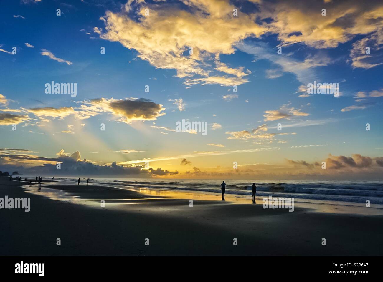 Ein bunter Sonnenaufgang am Strand mit Menschen zu Fuß entlang des Wassers im Sand Stockfoto