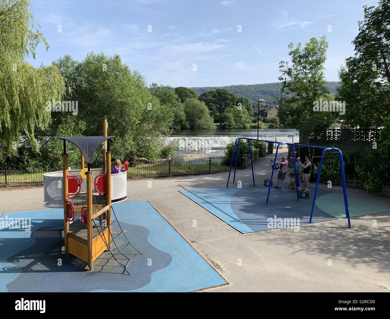 Kinderspielplatz in Otley, West Yorkshire, England. Ein oder zwei Kinder spielen vor der Fluss- und Wehr und Hügel dahinter. Stockfoto