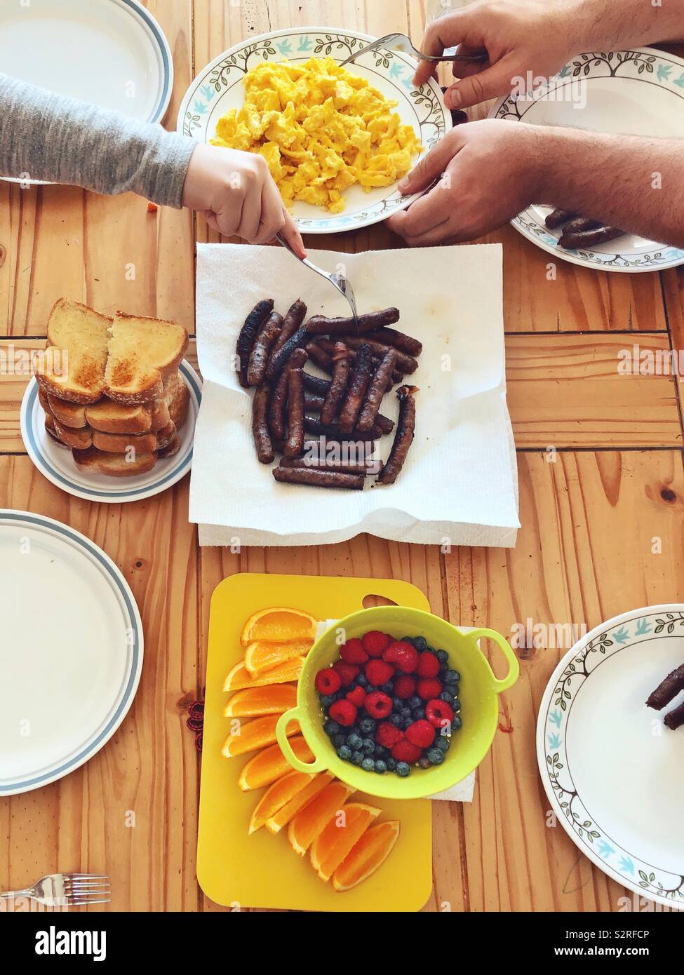 Ansicht der Hände mit Familie Frühstück mit Rührei, Würstchen und frischem Obst - Smartphone-aufgenommenes Stockfoto