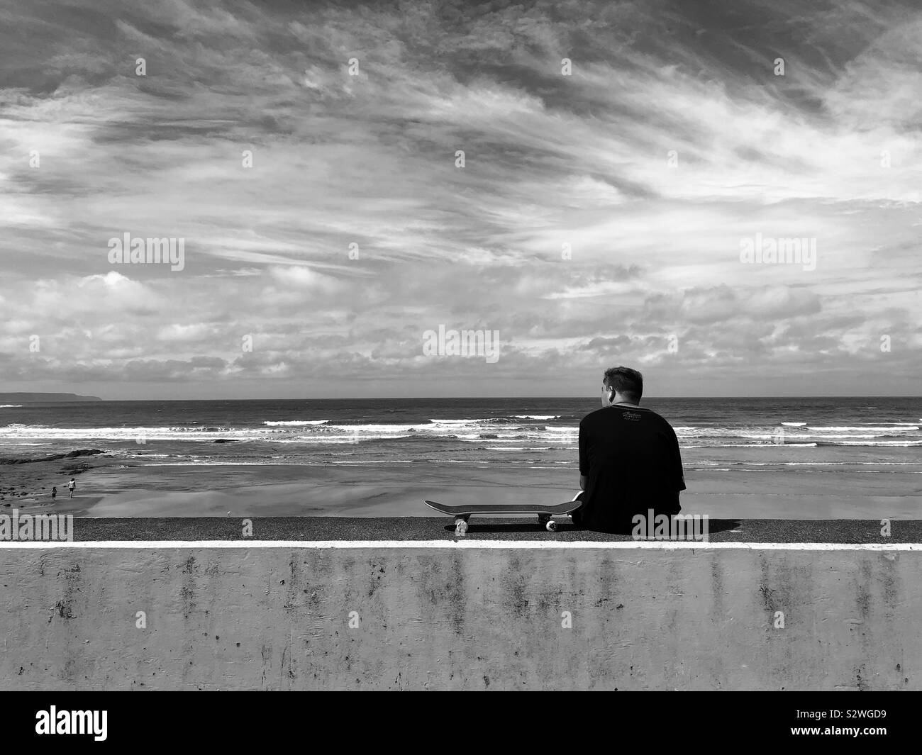 Junger Mann mit einem Skateboard sitzen auf einer ufermauer beobachten die Wellen an Westward Ho! Stockfoto