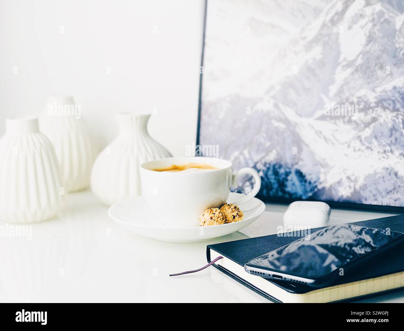 Aussicht auf Kaffee, Notebook, Handy, weiße Kopfhörer auf einem Sideboard mit Vasen und ein gerahmtes Bild eines Berges eingerichtet Stockfoto