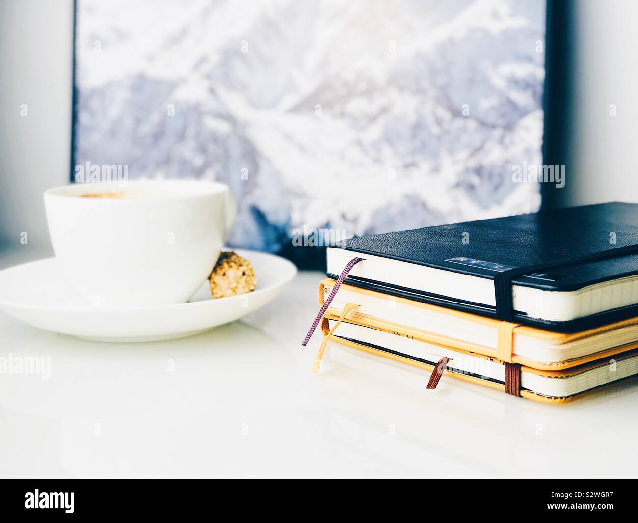 Close-up auf Heften von Notebooks, Kaffee und ein gerahmtes Bild von einem Berg in der Rückseite auf einem weißen oben auf einem Sideboard Stockfoto