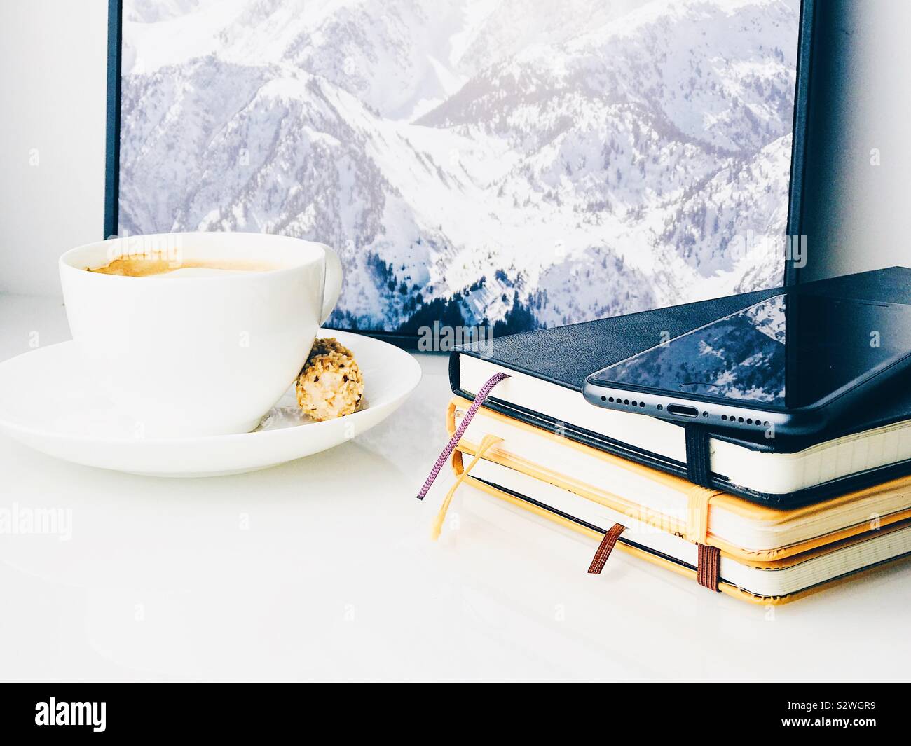 Close-up auf Heften von Notebooks mit einem Mobiltelefon, Kaffee und ein gerahmtes Bild von einem Berg in der Rückseite auf einem weißen oben auf einem Sideboard Stockfoto