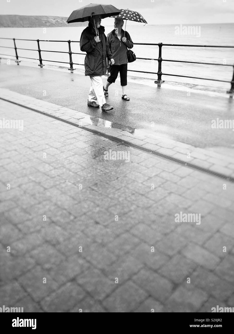Schwarz-weiß Foto eines Paares mit Sonnenschirmen zu Fuß entlang der Strandpromenade in der Regen Stockfoto