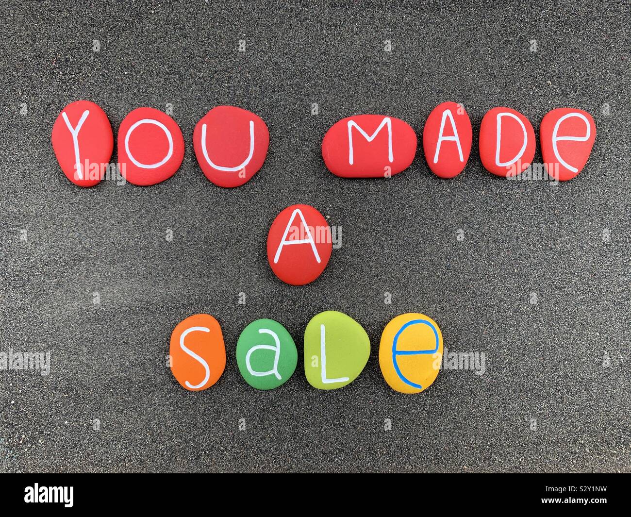 Sie haben einen Verkauf, iriginal commerce Schild mit einem creativecomposition von bunten und geschnitzte Steine über schwarzen vulkanischen Sand - Smartphone-aufgenommenes Stockfoto