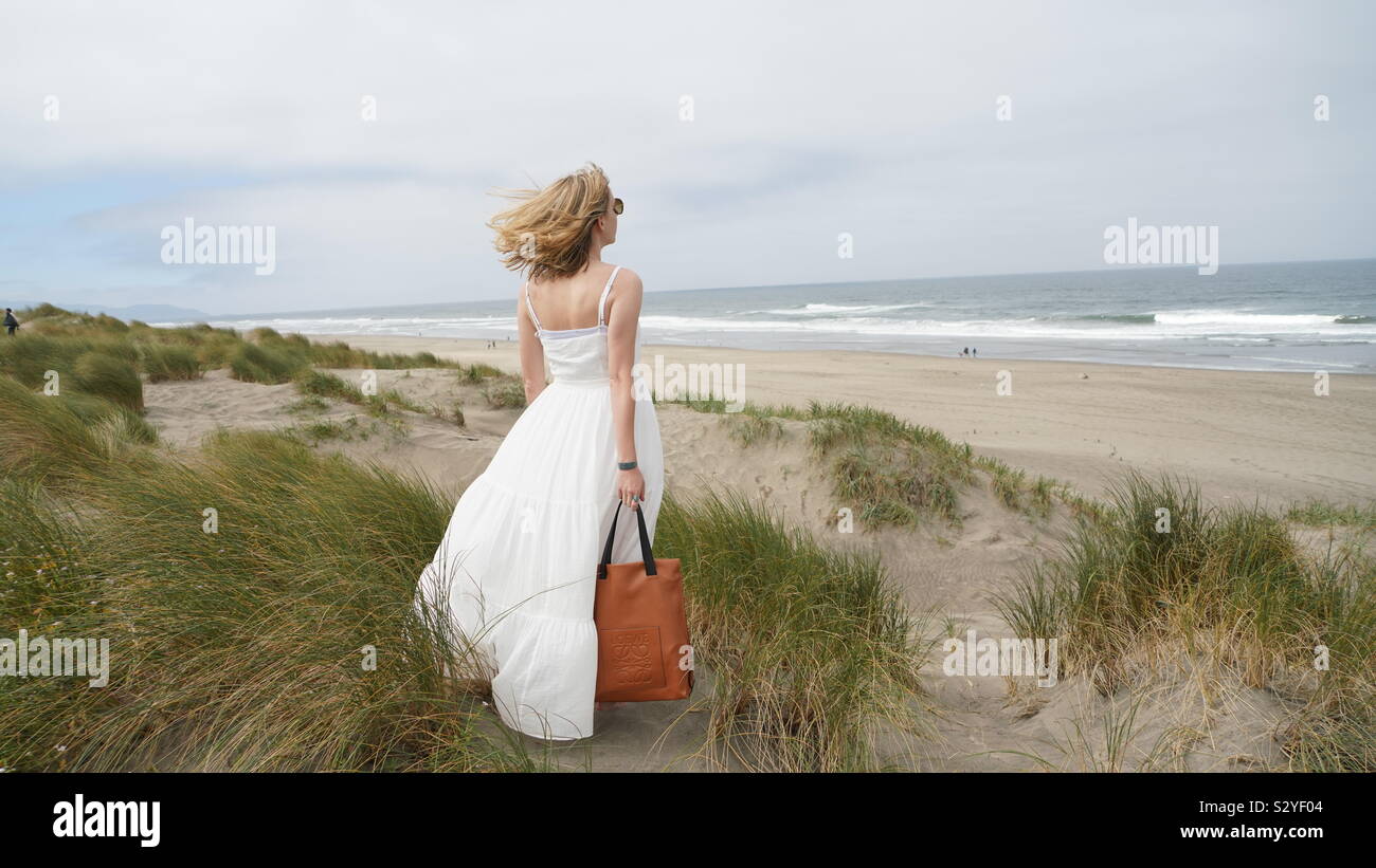 Junge blonde Frau, Haar weht im Wind, das Tragen von langen weißen Kleid, mit einem braunen Leder Tasche, mit Blick auf das Meer von einem sandigen Hügel Stockfoto