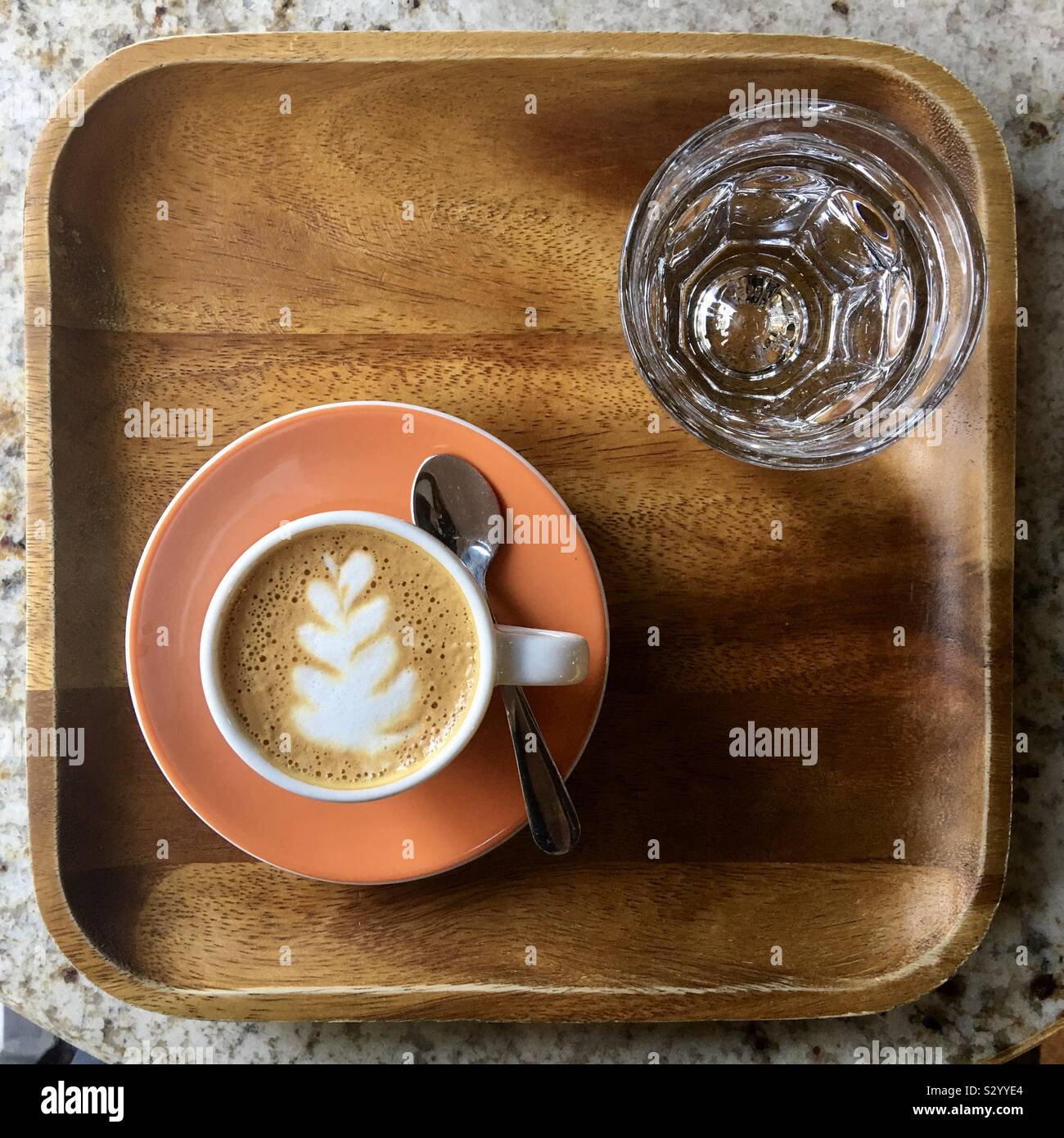 Tasse Espresso mit attraktivem Muster auf Schaum. Auf orange Untertasse serviert, auf kleinen hölzernen Fach mit einem Glas Wasser. - Smartphone-aufgenommenes Stockfoto