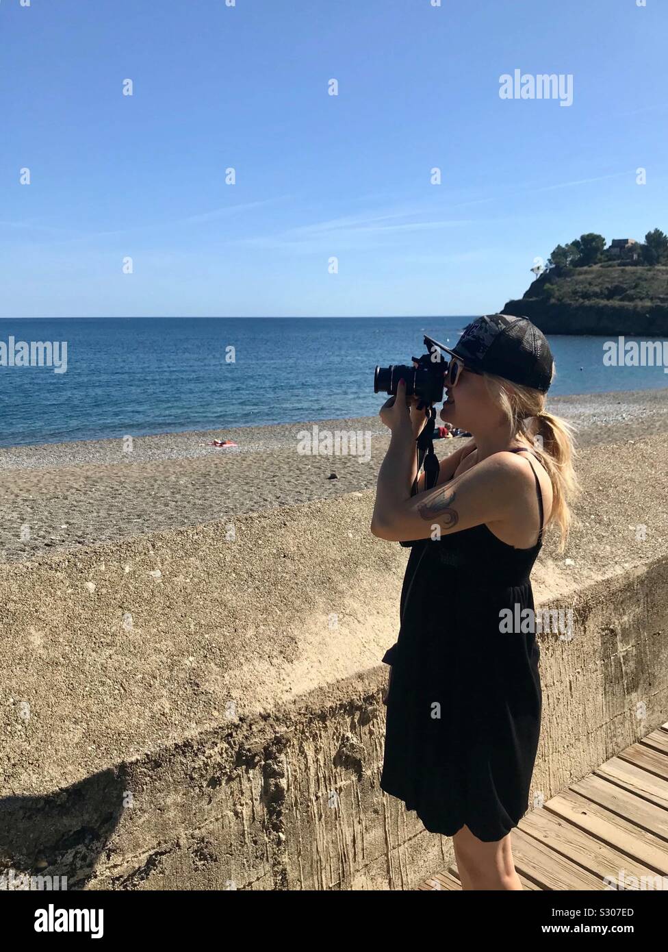 Blonde Mädchen mit Baseball Cap in Schwarz Sommer ärmelloses Kleid Fotos, die mit einer DSLR-Kamera am Meer. Weibliche Reise-fotograf. IG Stockfoto