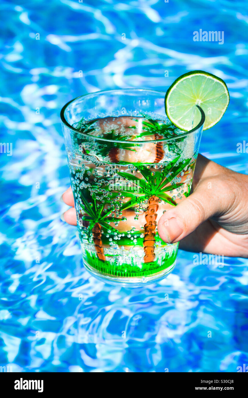 Hand mit einem Drink in einer Schale aus Kunststoff mit Palmen und einer Scheibe Limette, in einem Pool Stockfoto