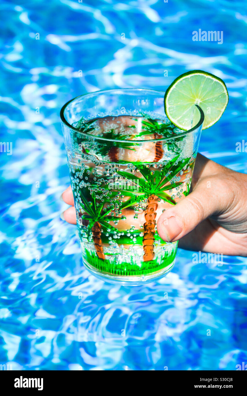 Hand mit einem Drink in einer Schale aus Kunststoff mit Palmen und einer Scheibe Limette, in einem Pool - Smartphone-aufgenommenes Stockfoto