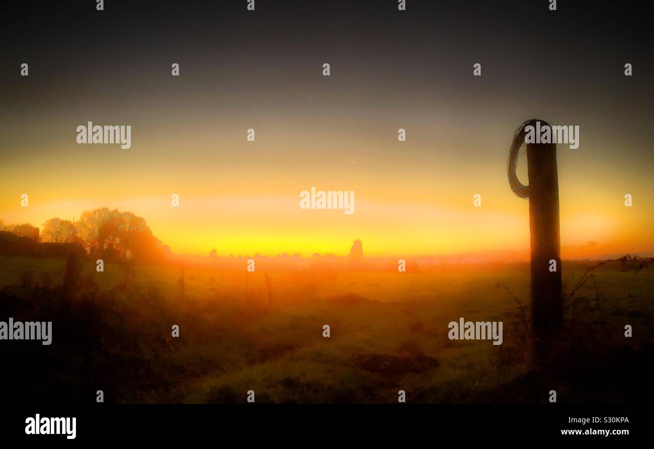 Feuriges rot orange leuchtende Sonnenaufgang oder Sonnenuntergang über einem Grasbewachsenen farmfield hinter einem Barb wired Post - Smartphone-aufgenommenes Stockfoto