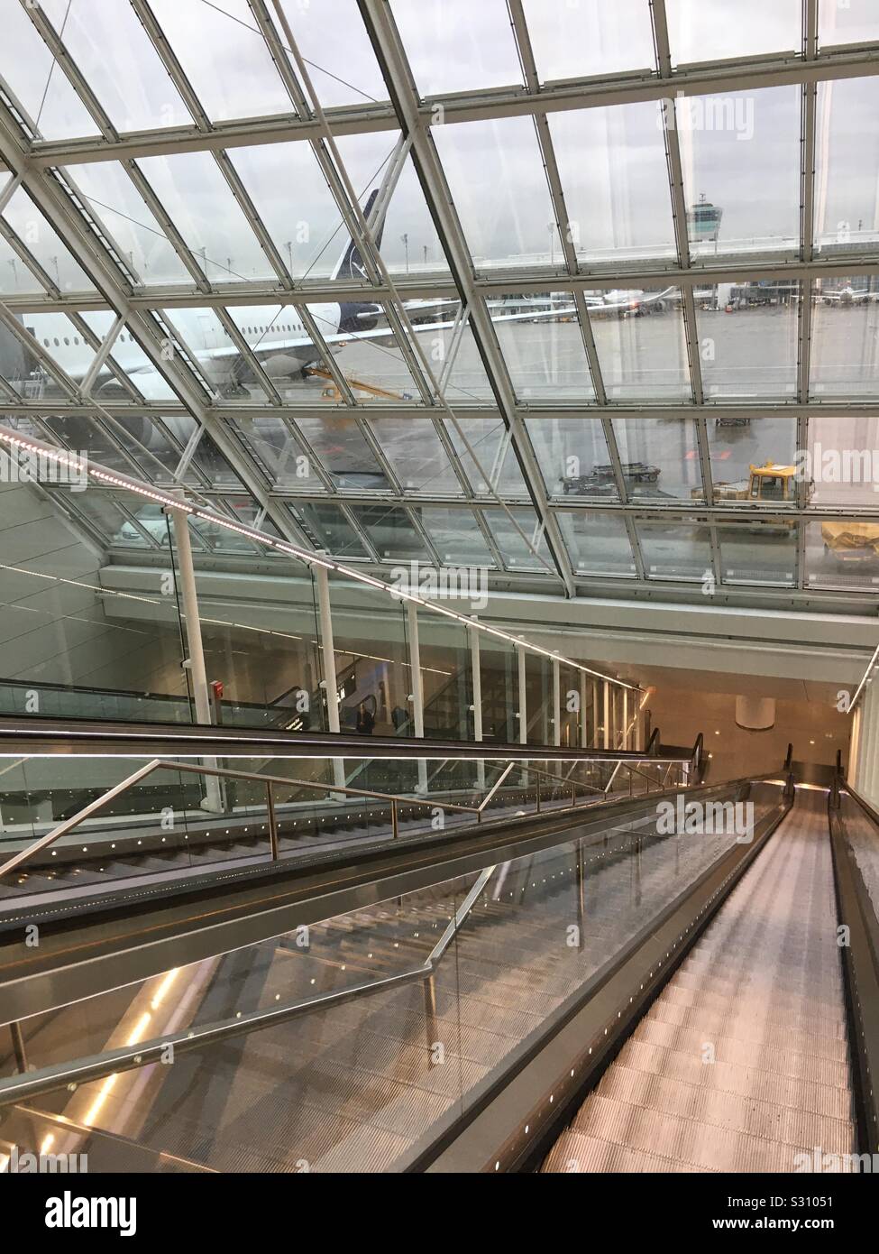 Nach oben und unten Rolltreppen am Flughafen München. Diesen Link das fahrerlose Shuttle für Transfers zu und von den T2-Satelliten. Das Foto zeigt auch ein Flugzeug am Boden, die gerade gewartet wird. Stockfoto