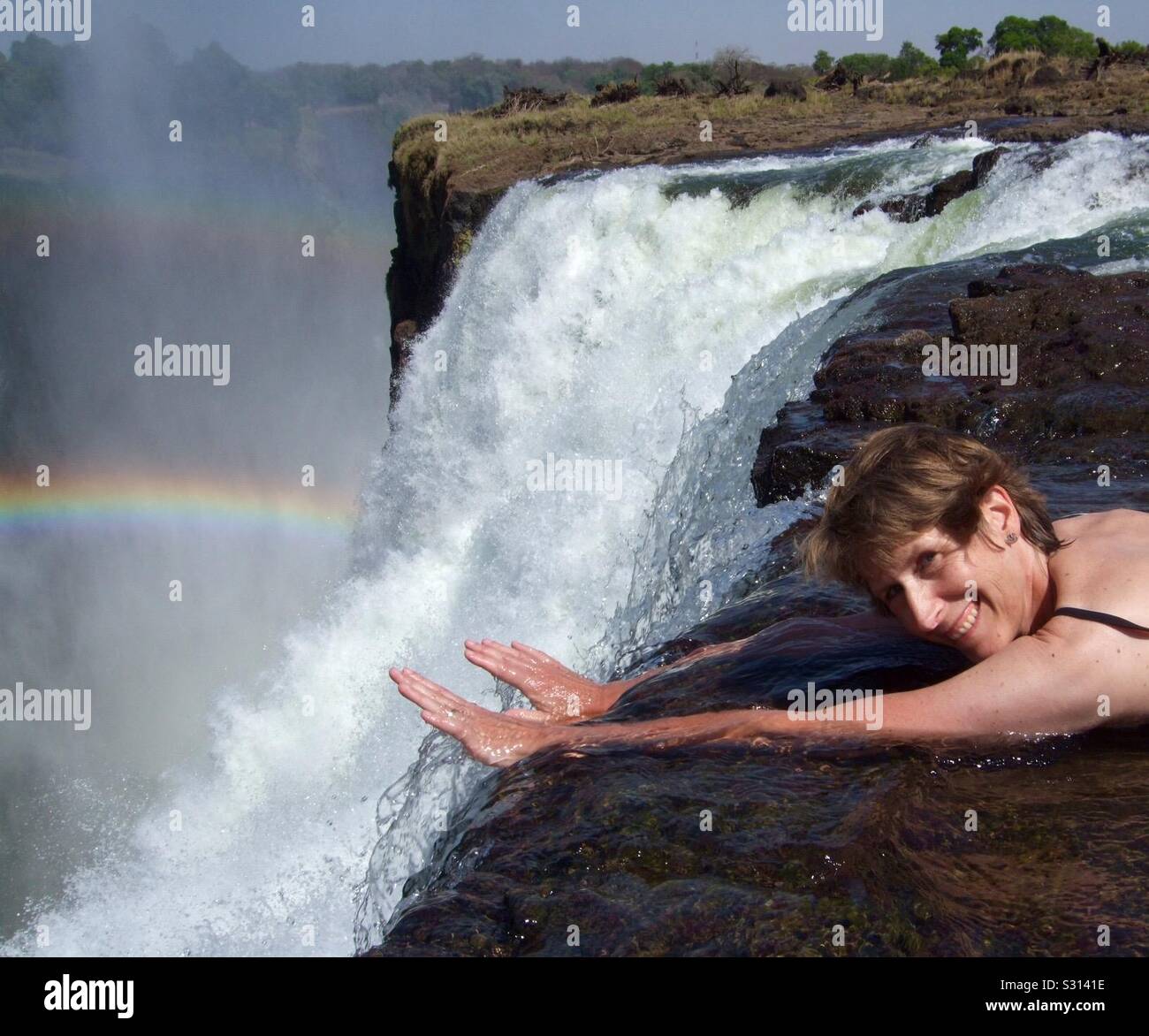 Frau in Badekleidung erreichen Sie über Victoria Falls bei Devil's Pool mit einem Regenbogen im Wasser Spray, Livingstone, Sambia Stockfoto