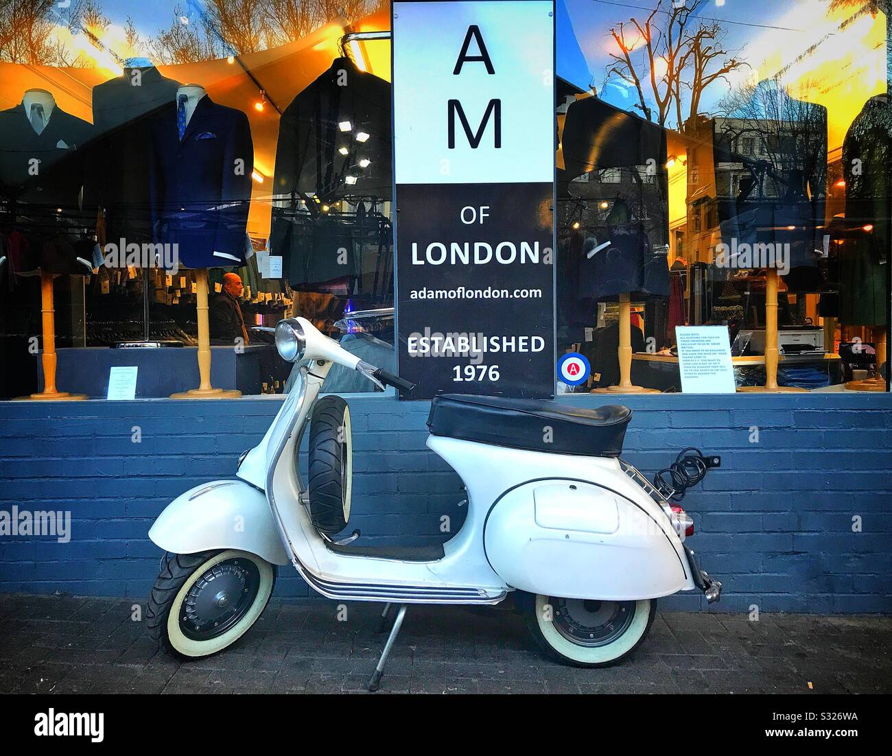 Auf dem Straßenbelag vor dem Schaufenster von Adam of London, einem Bekleidungshändler in der Portobello Road, Notting Hill, London, wird ein stilvoller Roller geparkt - Smartphone-aufgenommenes Stockfoto