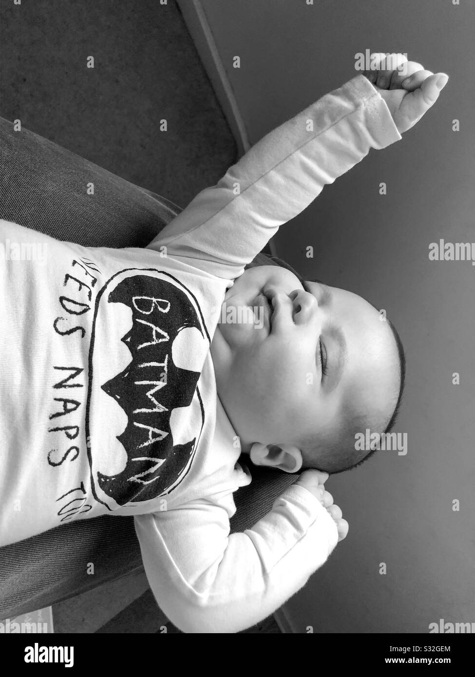 Ein 8-wöchiger alter Babyjunge, der auf dem Schoß seiner Väter nappt, während er als Batman verkleidet ist. Stockfoto