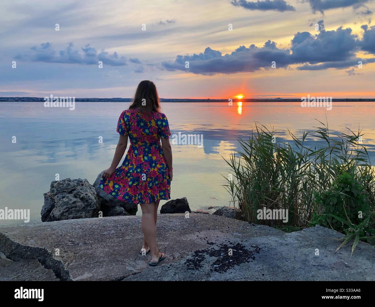 Frau im Kleid, die den Sonnenuntergang in der Nähe des Wassers beobachtet Stockfoto