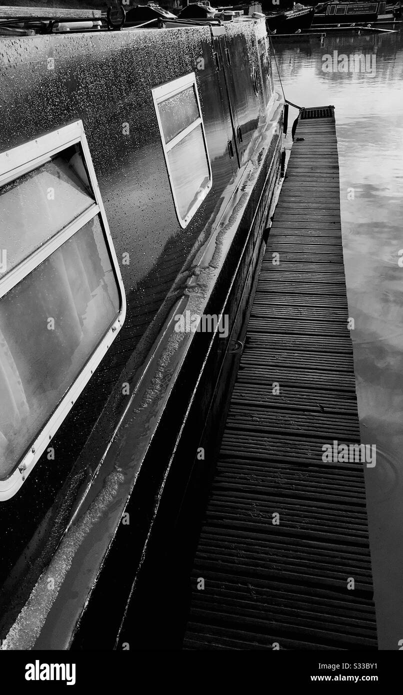 Mono-Bild, das einen Teil eines schmalen Bootes zeigt, das nach starkem Regen gegen einen Holzponton auf einem Yachthafen vermocht ist. Stockfoto