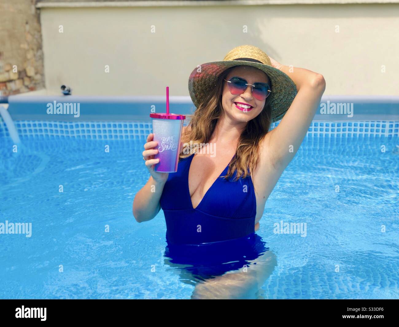 Frau mit blauem Bikini, Sonnenbrille und Strohhut, die einen Drink im Pool hält Stockfoto