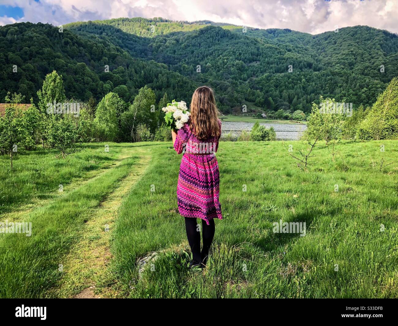 Frau in rosafarbenem Kleid, die einen Blumenstrauß aus weißem Flieder in den Bergen hält - Smartphone-aufgenommenes Stockfoto