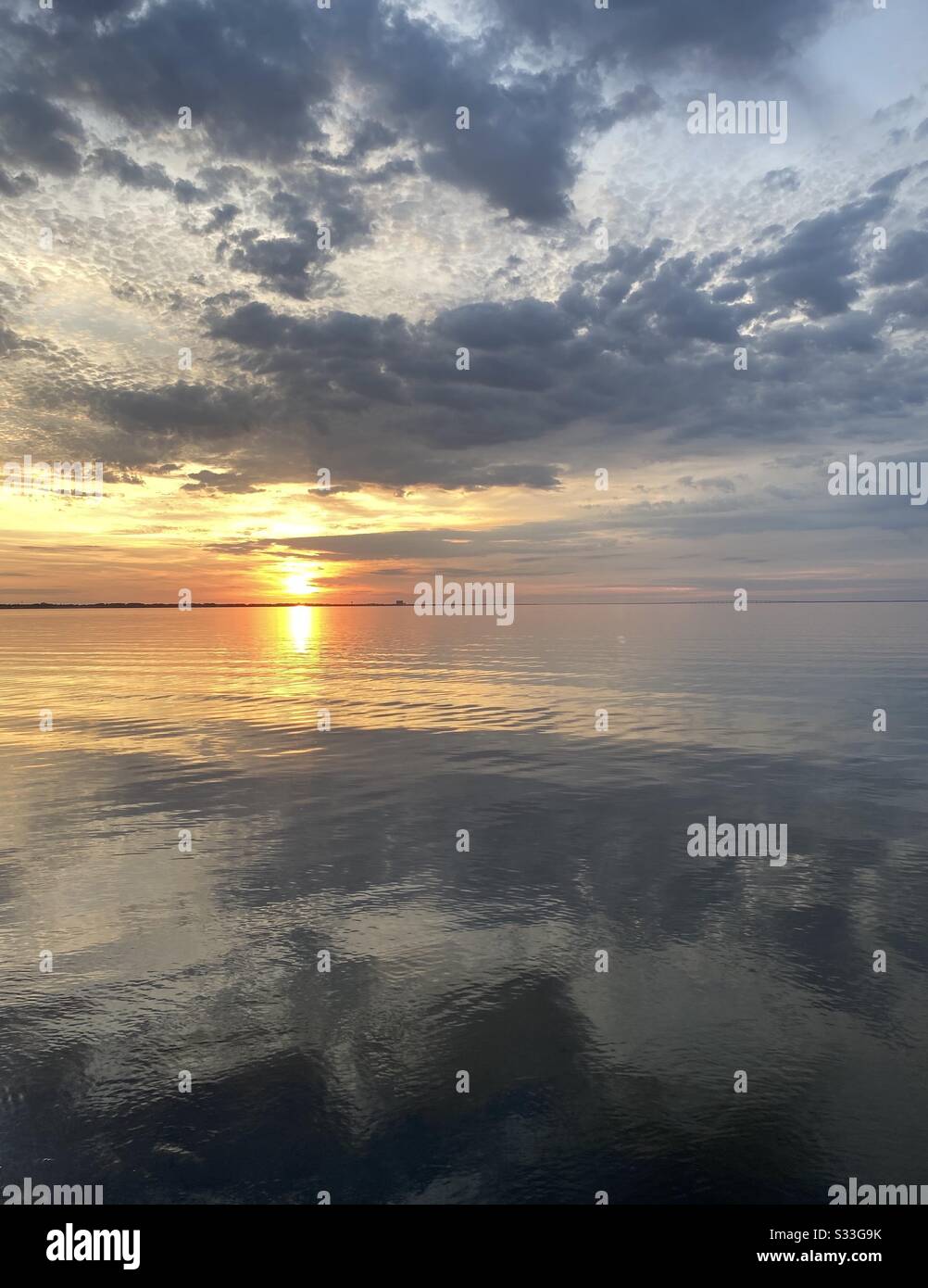 Goldener Sonnenuntergang über stillem Erkerwasser mit grauem bewölktem Himmel und Farbreflexionen auf dem Wasser - Smartphone-aufgenommenes Stockfoto