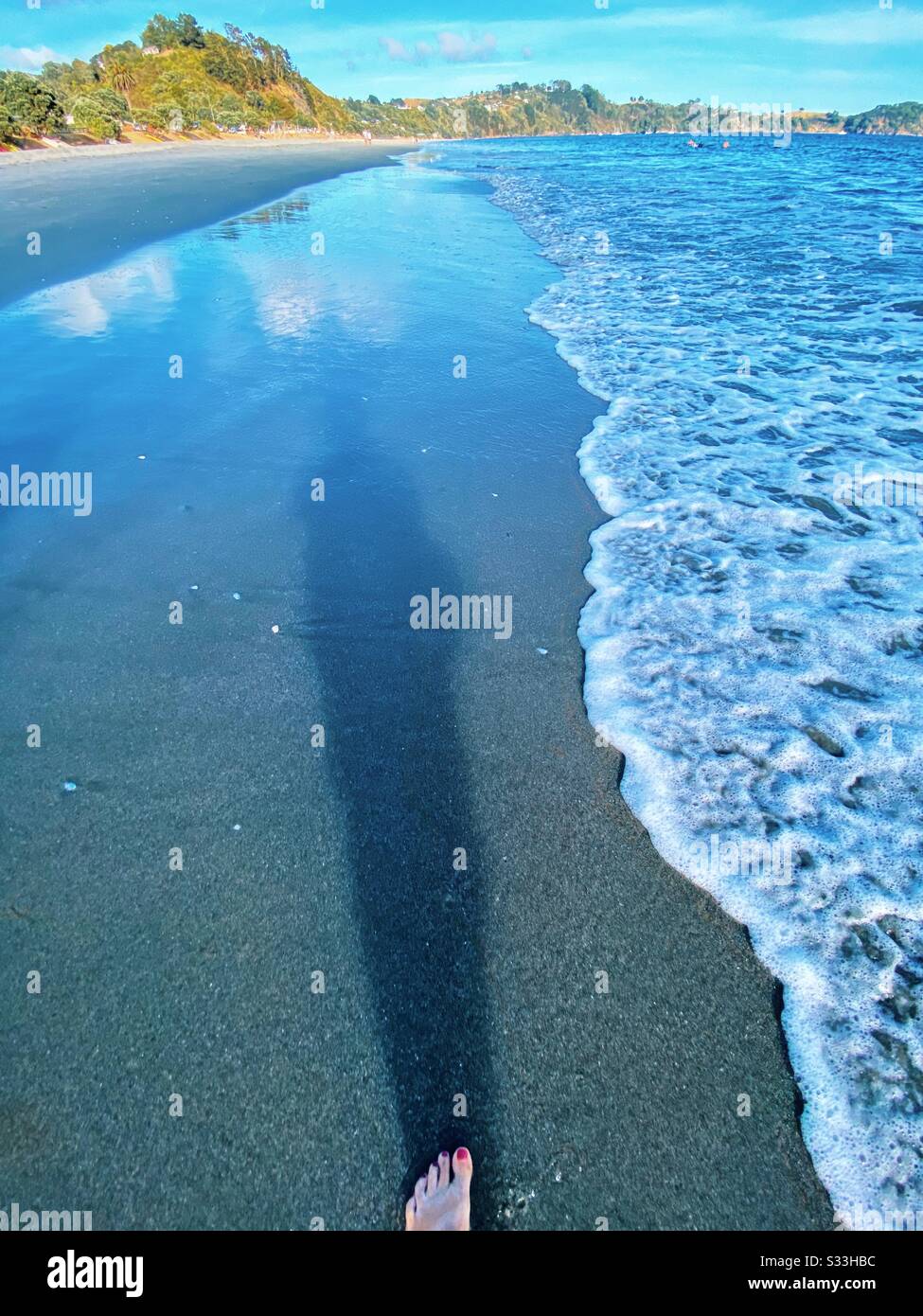 Ende des Fußes, der Zehen und des langen Schattens auf dem Sand, als die Wellen am Onetangi-Strand auf der insel waiheke in Neuseeland auf das Ufer schlugen. Morgen im Februar 2020. Stockfoto