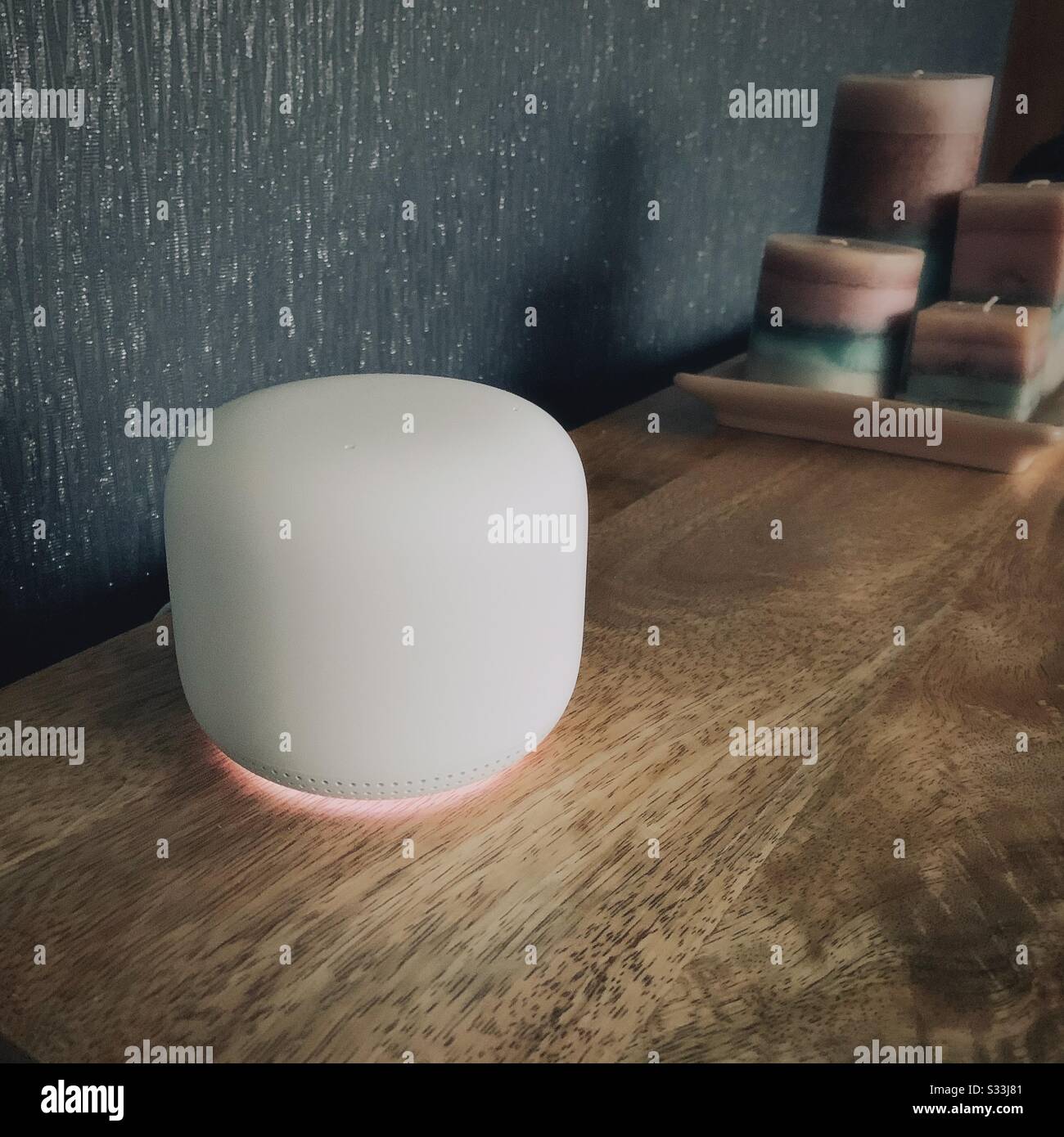 Nahaufnahme eines Goggle Nest-Heimrouter-WiFi-Punktes mit leuchtenden Lichtern. Moderne Technologie zur Schaffung eines Netzes von Breitbandnetzwerken. Verschwommene Kerzen im Hintergrund auf Holzregal Stockfoto