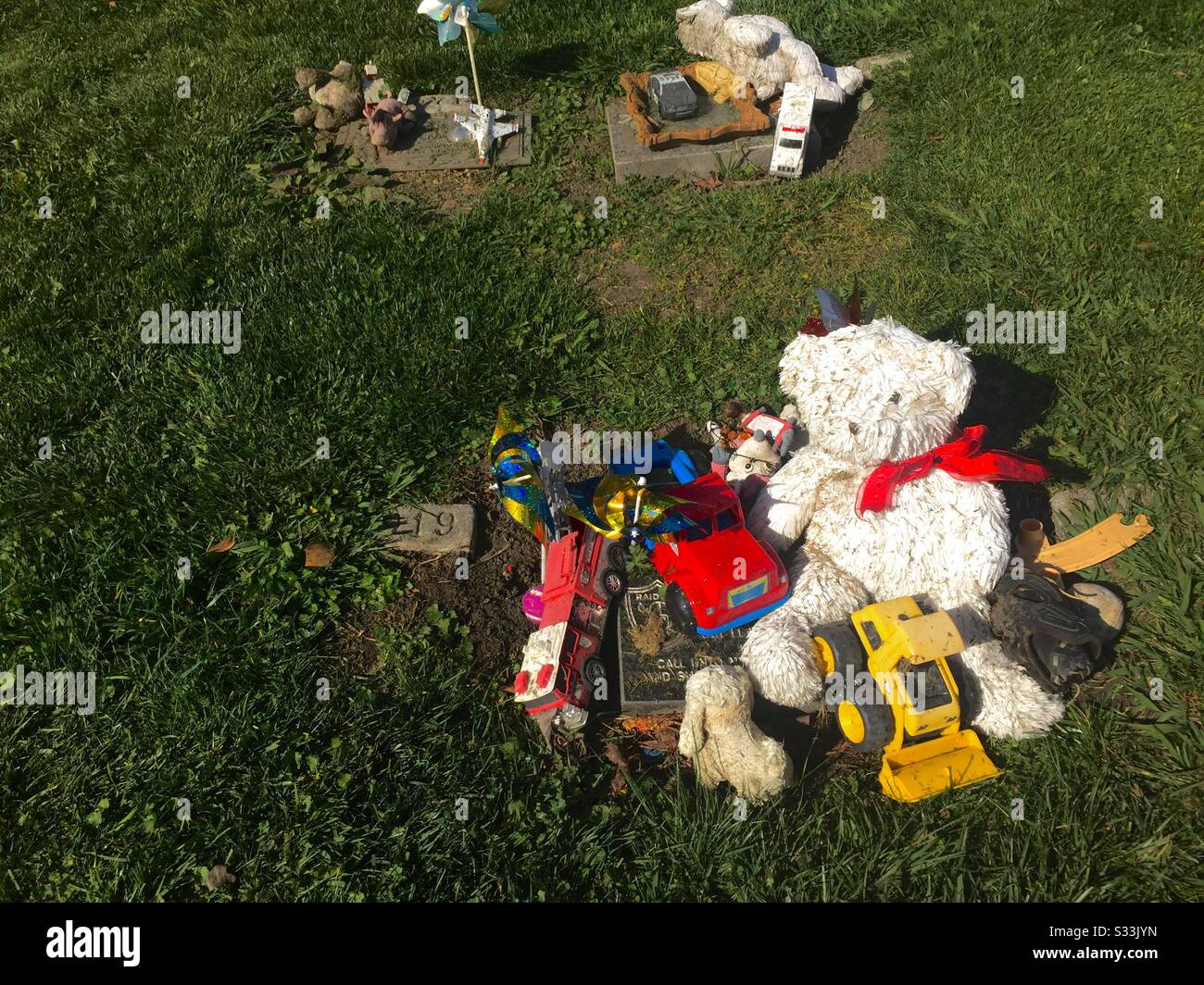 Kindergräber mit verwittertem Spielzeug - Plastikwagen, weißer Teddybär mit rotem Bogen, gefüllte Tiere - auf Grabsteinen aufgestapelt. Friedhof Mit Blick Auf Die Berge. Oakland, Kalifornien. - Smartphone-aufgenommenes Stockfoto