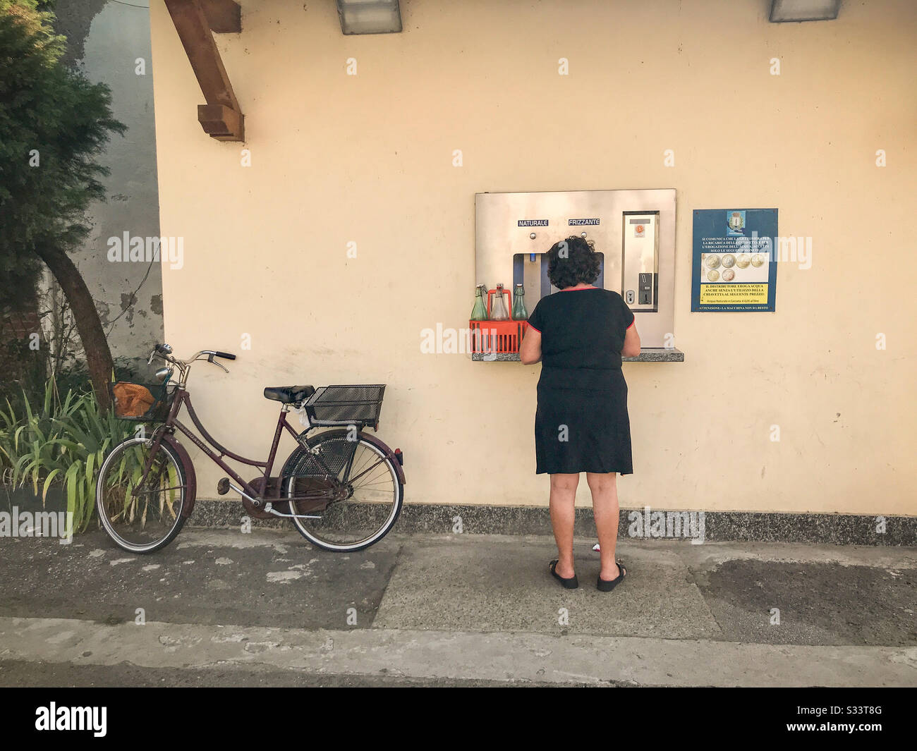 Der Rücken einer Frau, die mit ihrem Korb mit Glaswasserflaschen im ländlichen Italien an einer Wasserauffüllstation steht. Ihr Fahrrad ruht auf dem Gebäude. Stockfoto