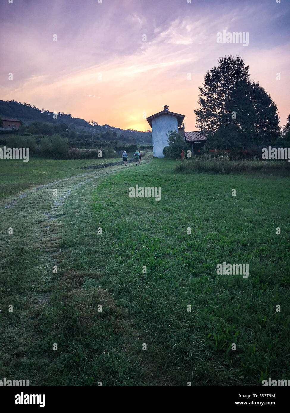 Romantische Landschaft bei Sonnenaufgang mit Rucksacktouristen, einem alten weißen Gebäude, Pfad und üppiger grüner Landschaftsgestaltung. Stockfoto