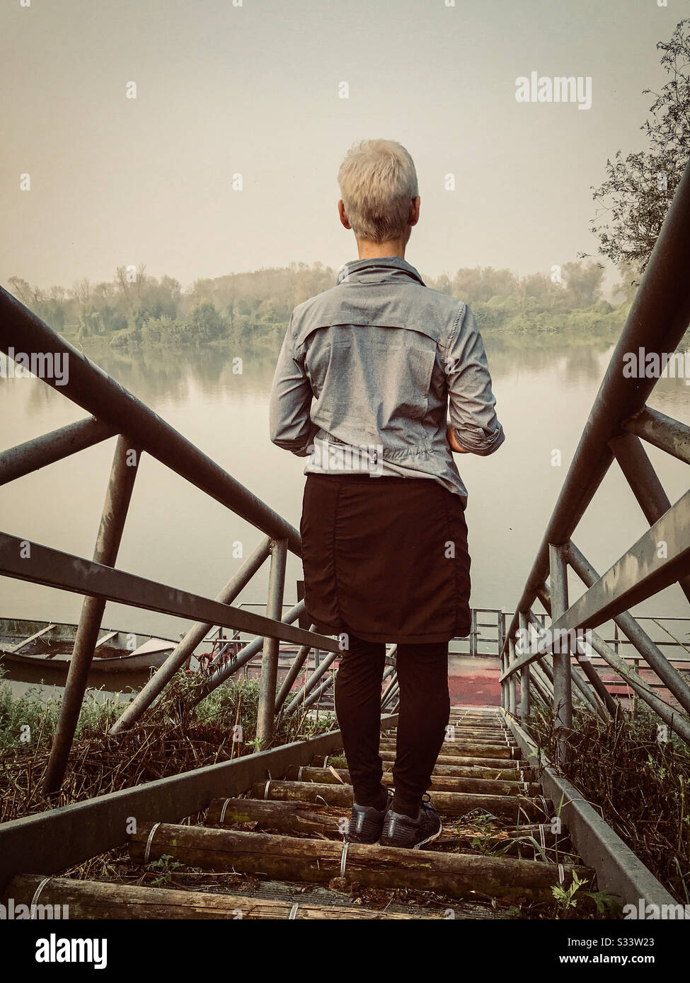Vintage-aussehendes Foto einer Frau, die ein graues Hemd und einen schwarzen Rock trägt, der auf einer Treppe steht, die in Italien zu einem Fluss führt. - Smartphone-aufgenommenes Stockfoto