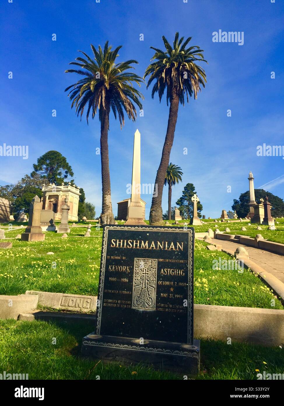 Weißer Obelisk-Grabmarker zwischen zwei Palmen. Kopfstein mit Namen Shishmanian, Kevork, Astghig. Gruft, Gräber, Gräber. Mountain View Cemetery, Oakland, Kalifornien. Stockfoto