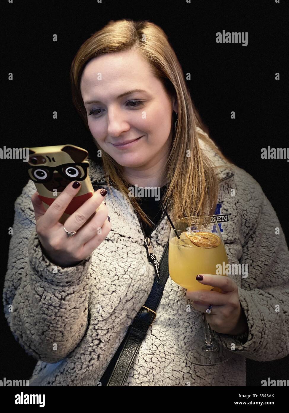 Isolierte kaukasische junge Frau vor schwarzem Hintergrund, die ihr Handy in der einen Hand betrachtet und ein alkoholisches Getränk in der anderen hält, während sie lächelt. - Smartphone-aufgenommenes Stockfoto