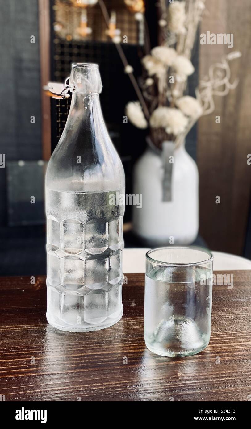 Einfache Glasflasche mit Wasser neben einem Glas Wasser auf einem Tisch mit einer Vase von Blumen im Hintergrund. Stockfoto