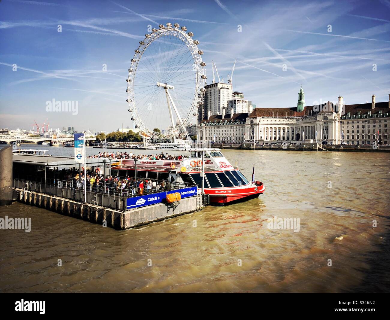 Am Westminster Pier auf der Themse ist ein Stadtkreuzfahrtschiff zu sehen, mit Blick auf das Millennium Wheel und die Flugzeugkontrails in London Stockfoto