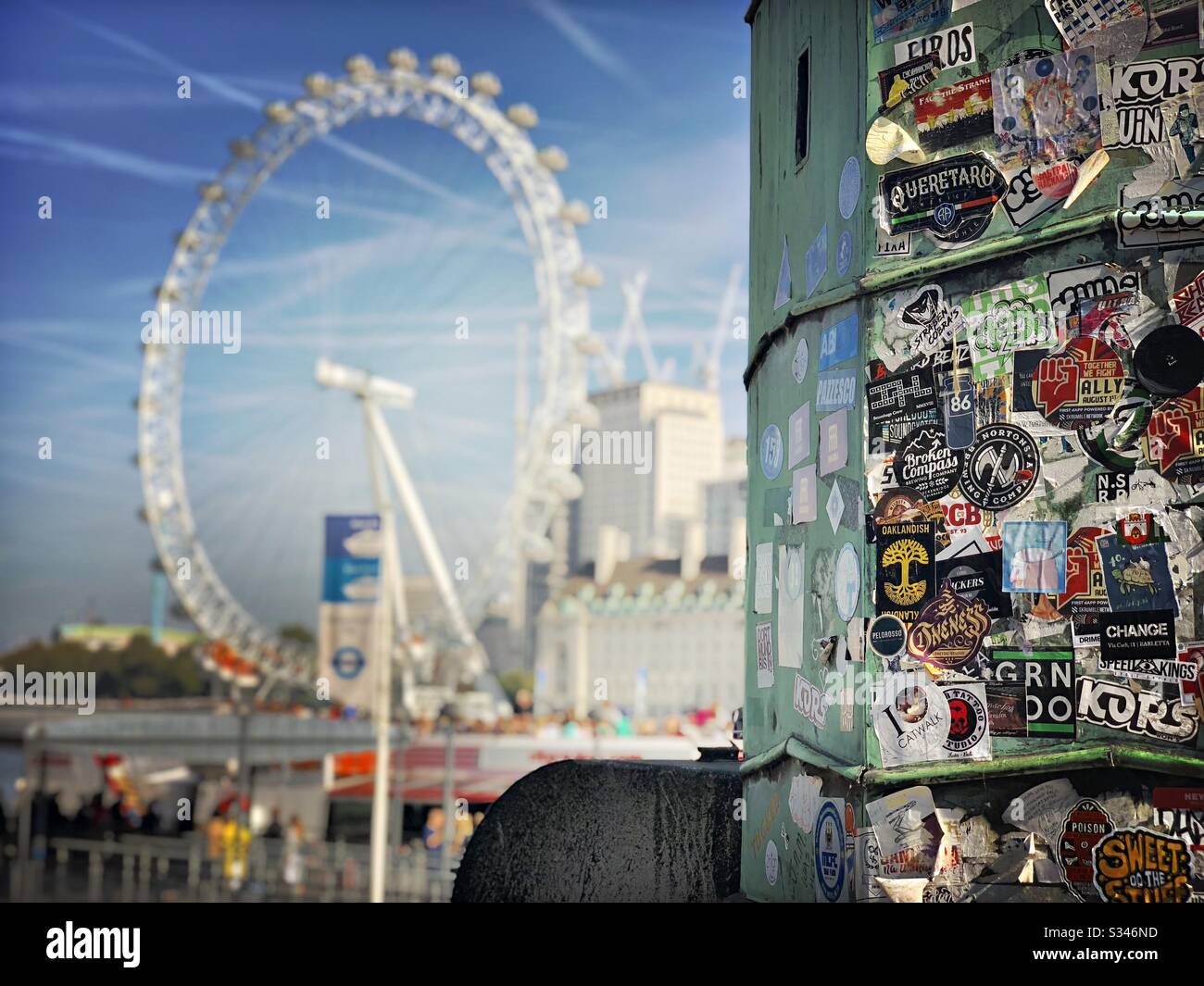 Nahansicht eines mit Touristenaufklebern bedeckten Lampenpostens am Westminster Pier mit verschwommener Sicht auf das London Eye/Millennium Wheel - Smartphone-aufgenommenes Stockfoto