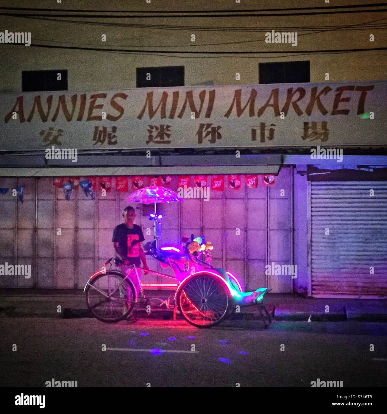 Ein Trishaw, vor Ort bekannt als "Beca", dekoriert mit LED-Lampen, die auf dem Nachtmarkt in Batu Ferringhi, Penang, Malaysia auf Geschäfte von Touristen warten - Smartphone-aufgenommenes Stockfoto