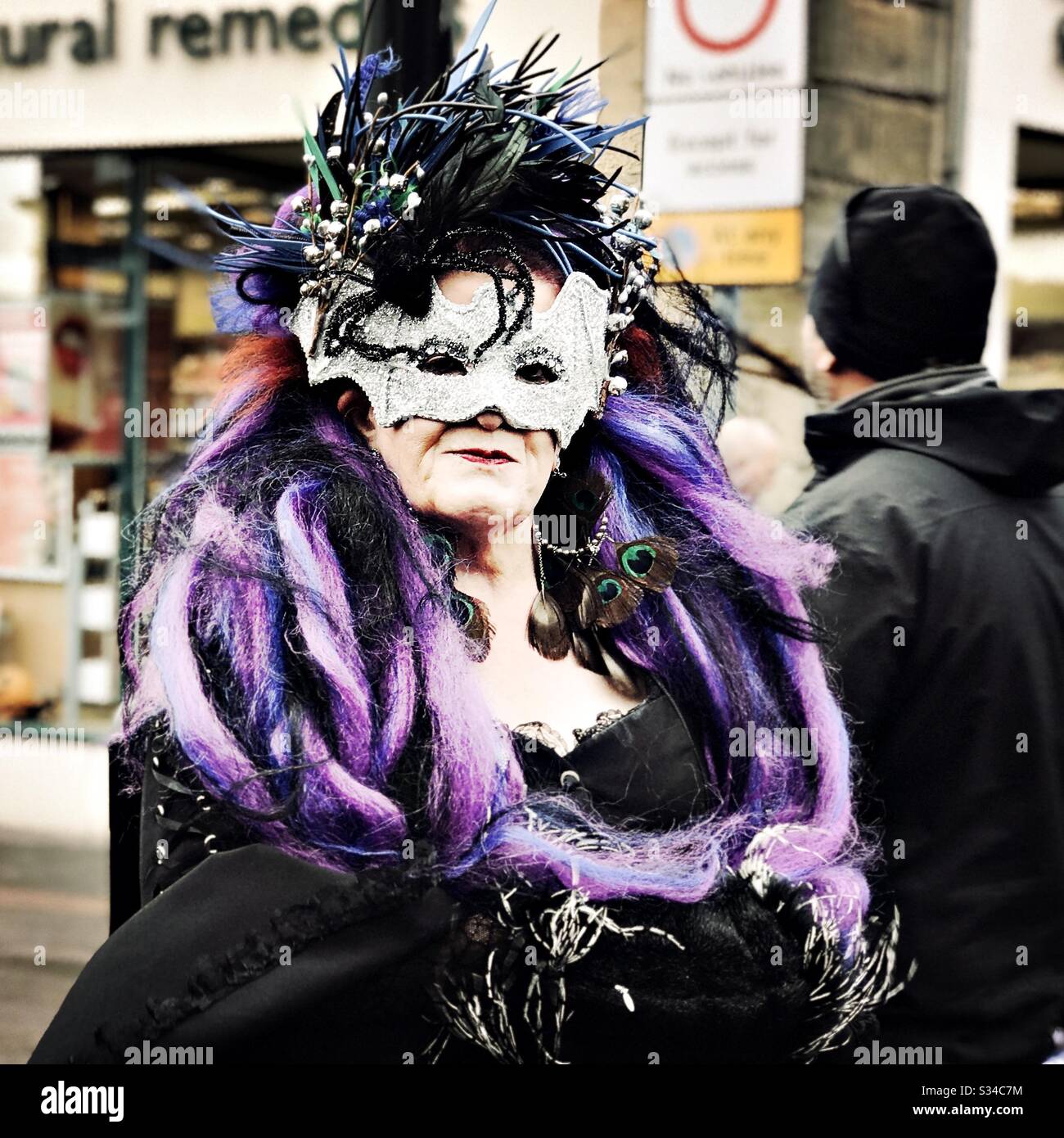 Person in einem kreativen Kostüm während einer Cosplay-Veranstaltung. Whitby Goth Weekend. Selbstbewusste ältere Frau in Maske und violetter Boa - Smartphone-aufgenommenes Stockfoto