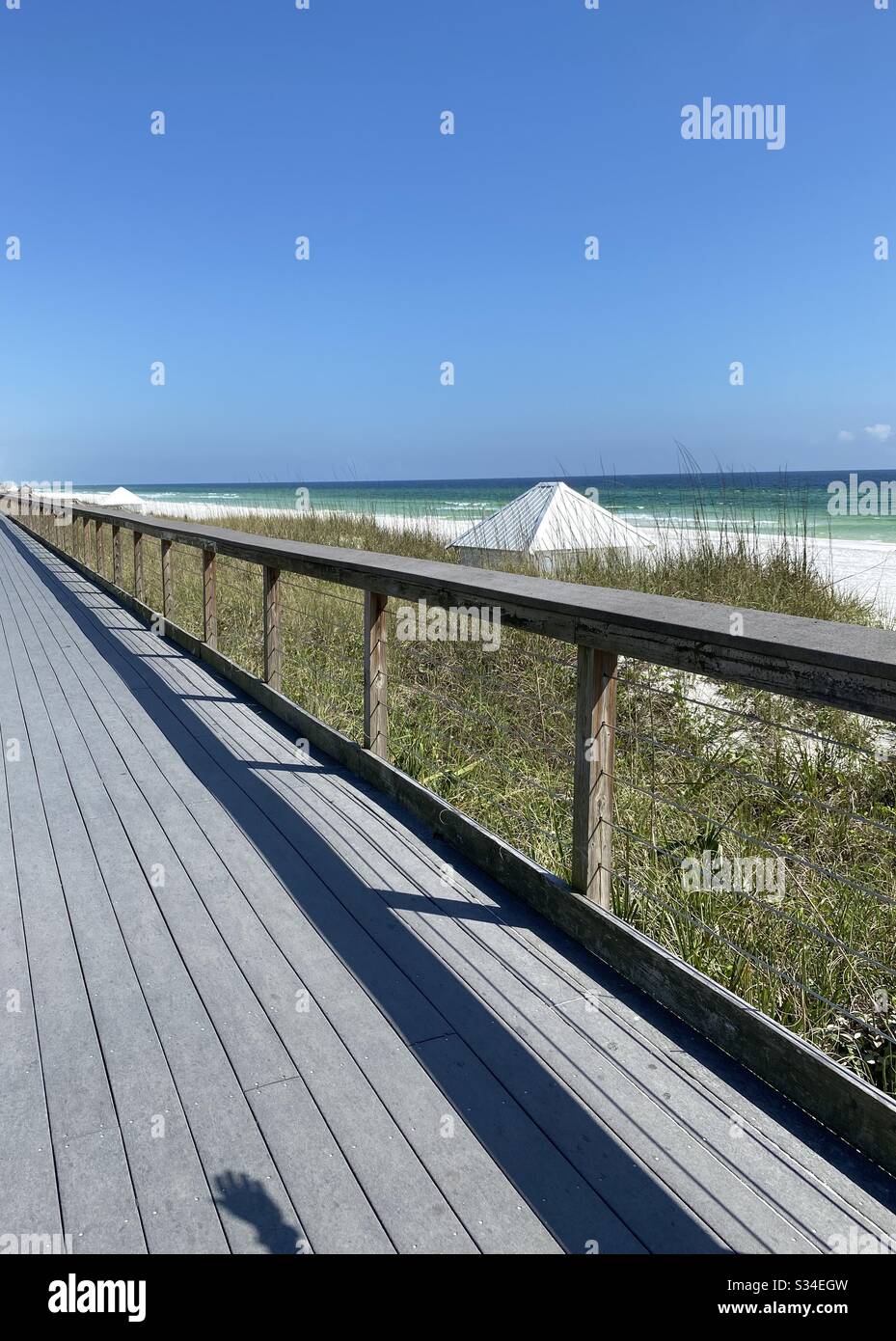 Malerischer Gehweg entlang des smaragdgrünen Golfs von Mexiko Wasser und weißer Sandstrand mit einem Schatten von einer Hand auf dem Gehweg - Smartphone-aufgenommenes Stockfoto
