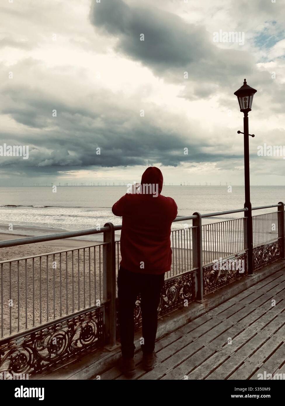 Ein junger Mann in einem roten Hoodie, der auf dem Skegness Pier steht, macht ein Foto von einem Sturm auf das Meer Stockfoto